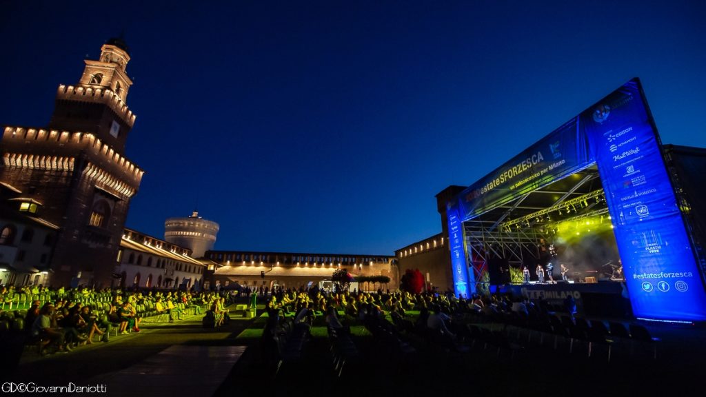 Nella notte illuminata, sulla destra il castello sforzesco di milano e sulla sinistra il palco illuminato di giallo e blu. in mezzo la platea con il pubblico seduto