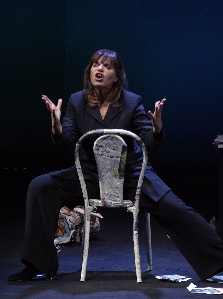 Civitafestival - Elena Bonelli seduta a cavalcini su una sedia messa al contrario, indissa un completo di giacca e pantaline blu, sta recitando con le braccia aperte e le mani tese