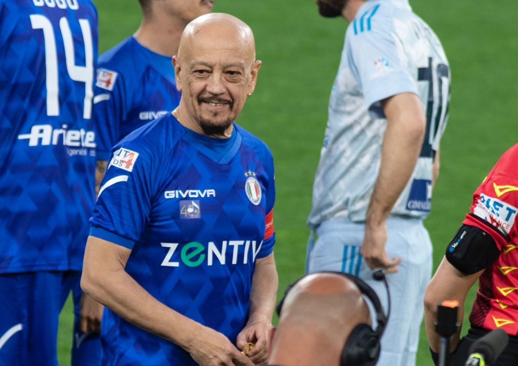 Enrico Ruggeri con la maglia azzurra della nazionale cantanti, sorride, in campo durante una partita della squadra dei cantanti. Nella foto anche altri giocatori di cui non si vede il viso, uno vestito di bianco, l altro di azzurro. In basso spunta la testa calva di un cronista che ha le cuffie d'ascolto in testa