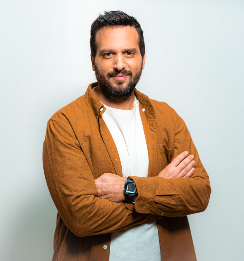 Gaetano Pecoraro con capelli neri e barba nera, incrocia le braccia, indossa t shirt bianca e camicia arancione