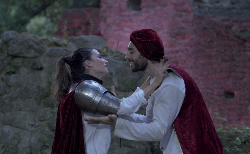 Una ragazza con armatura cavalleresca, con lunghi capelli newri legati in una coda di cavallo abbraccia un uomo vestito di bianco con un turbante rosso