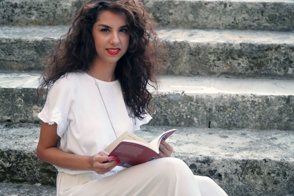 Mariagela Maio seduta su dei gradini di pietra, indossa un vestito bianco girocollo con maniche corte, guarda verso l'obiettivo sorridente e tiene in mano il suo libro