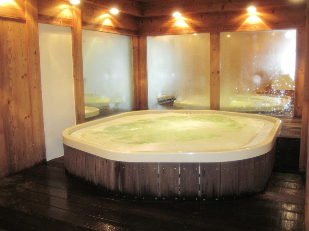 Una sala di un centro termale con una vasca gigante idromassaggio al centro e tutto intorno delle porte di legno e vetro trasparente delle cabine sauna