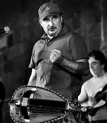 Lou Dalfin foto in bianco e nero con un uomo con t shirt e berreto con la mano appoggiata sul petto. Vicino a lui un altro ragazzo e uno strumento a corde siumile a un mandolino