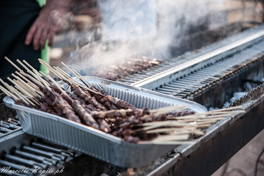 degli arrosticini di pecora in una teglia retangolare di alluminio appoggiata su una griglia dove si stanno cuocendo altri arrosticini