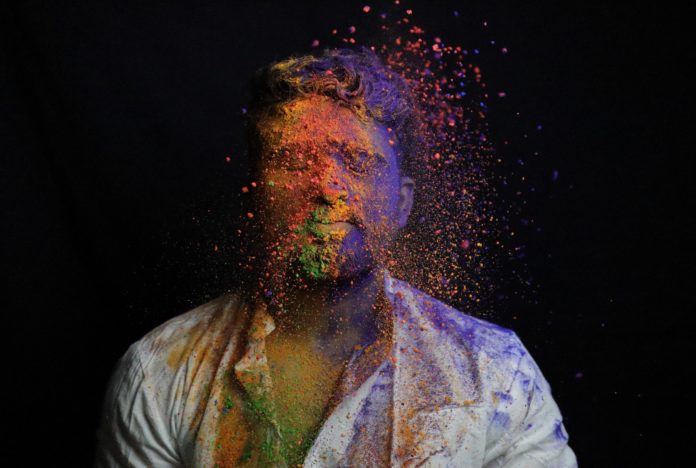 karel Music Expo. nella foto un uomo con gli occhi chiusi viene spruzzato da una polvere di tuti i colori sul volti e sul petto.