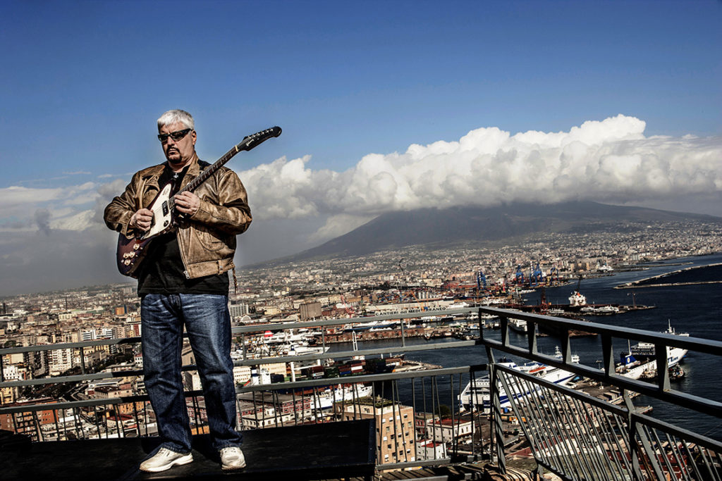 Una veduta panoramica di Napoli e Pino Daniele sulla destra, concapelli bianchi, vestito con jeans e giubbotto beige, mentre suona la chitarra