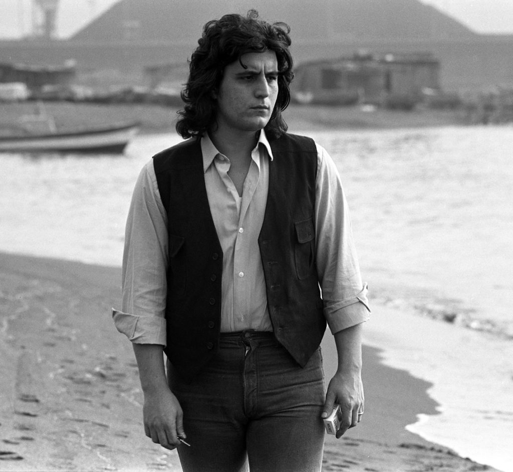 Pino Daniele in riva al mare, passeggia assorto, indossa pantaloni scuri, gillè e camica. foto in bianco e nero