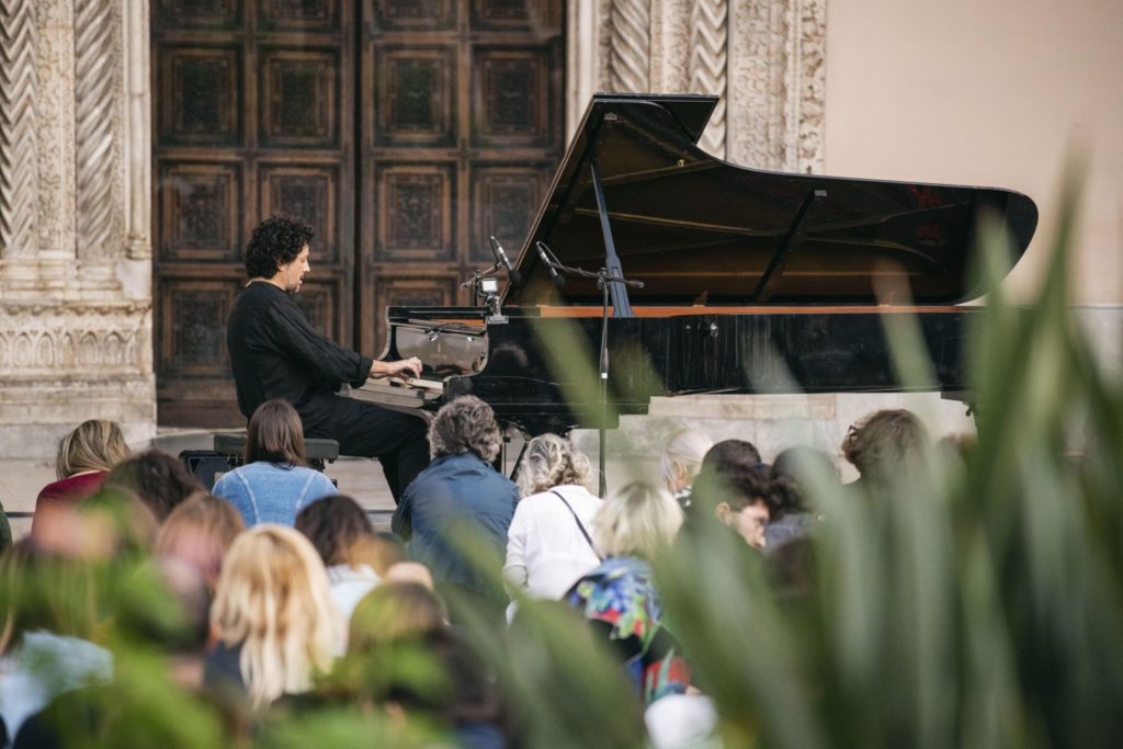 Un ragazo con capelli lunghi sulle spalle riccioluti, suona il pianoforte in piazza davanti a tanta gente