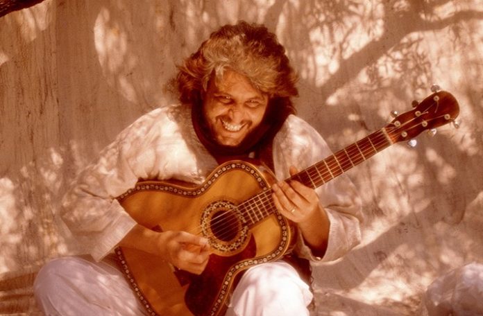 Pino Daniele seduto con la chitarra, vestito di bianco, ride. La foto è tutta in tinta seppia