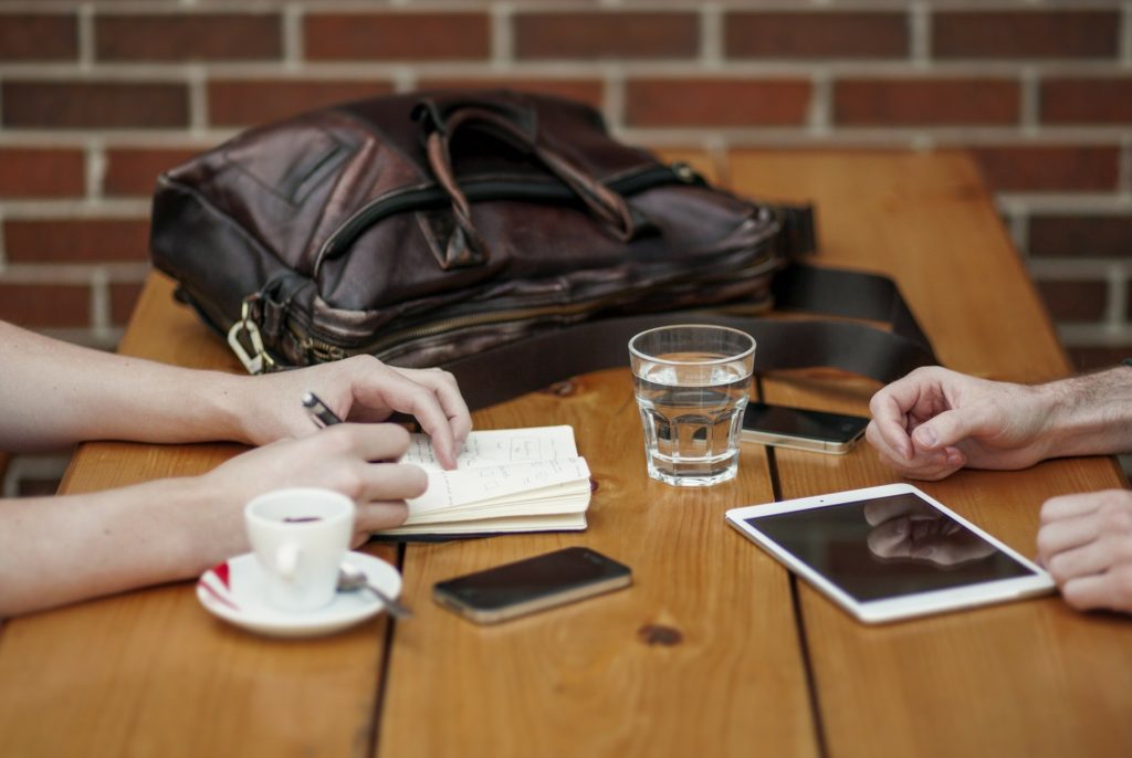 un tavolo con sopra una vboretta grande da donna, una tazza di caffè, un bicchiere d'acqua e due persone sedute. Non si vedono i volti ma solo le mani che scrollano il vetro del cellulare