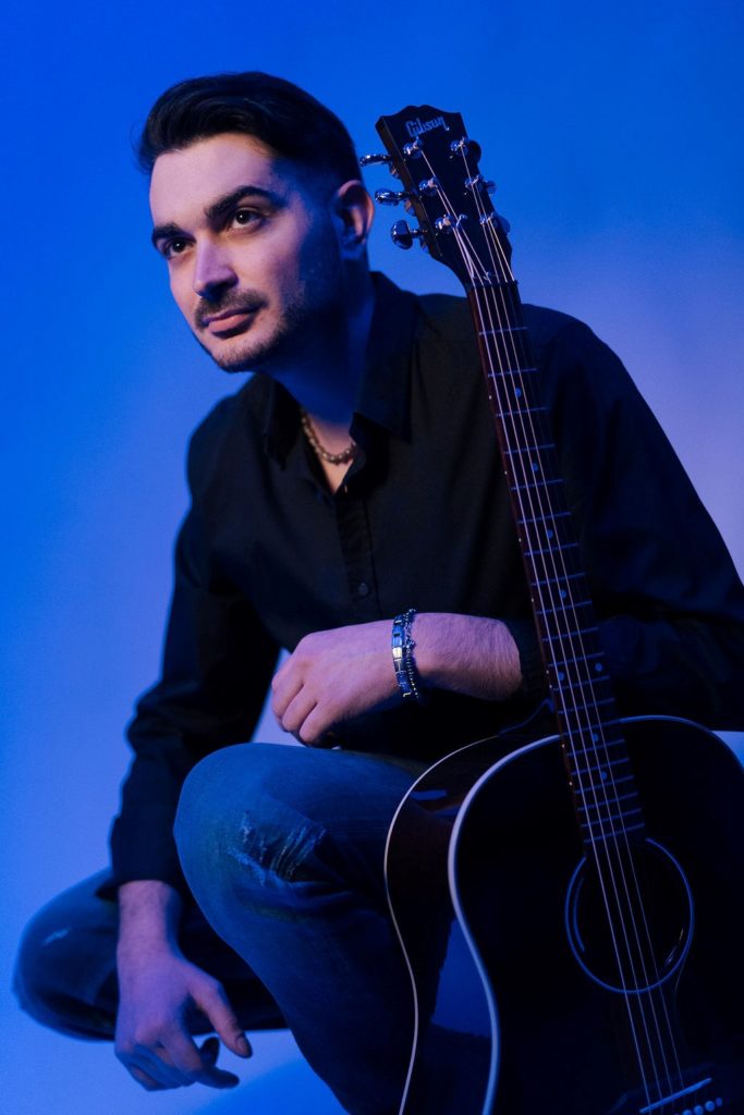 gianluca centenaro, indossa jeans e camicia nera, inginocchiato con la chitarra a tracolla