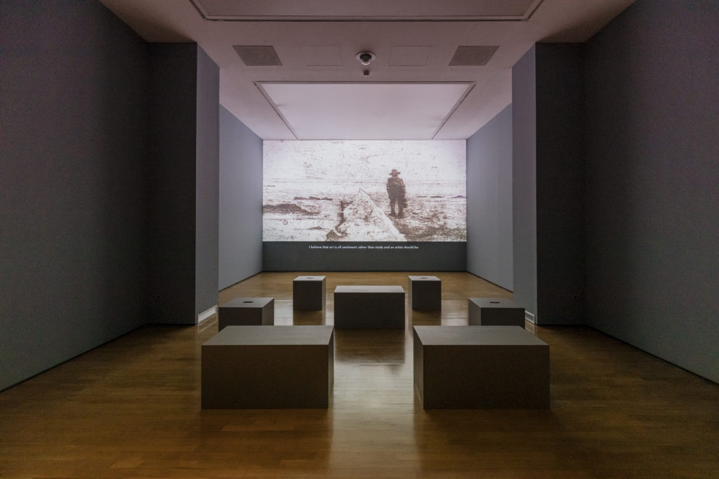 Giovsanni FAttori - una sala della msotra con una immagine proiettata su uno schermo, in bianco e nero, con il disegno di un uomo di schiena in mezzo a terra secca