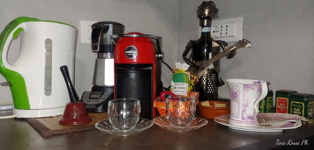 un angolo di cucina predisposto per il caffè con una caffettiera elettrica moka, una espresso, un bollitore bianco e verde, due tazizne da caffè in vetro con piattino, un cestello piccolo con delle cialde, una tazza da te rosa con un piattino rosa e bianco allungato, dietro tutto un porta bottiglia fatto a forma di persona che suona la chitarra