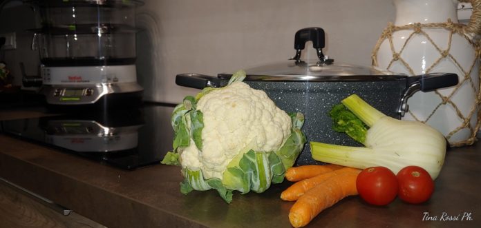 Cucina innovativa - nella foto in primo piano un cavolfiore, un finocchio delle carote e due pomodorini sono appoggiati su un piano di una cucina. Dietro di loro una pentola in pietra grigia e sullo sfondo un piano a induzione in vetroceramica nero e una vaporiera