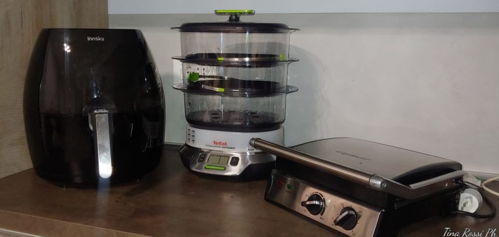 sul piano di lavoro di una cucina ci sono un friggitrice elettrica ad aria nera, una vaporiera con dei cestelli traspareenti e una piastra tostiera