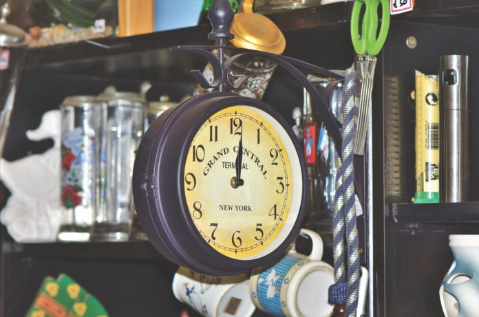 un orologio analogico che segna mezzogiorno, con il quadrante rotondo in legno, appeso in mezzo a vecchi oggettii in un mercatino dell'usato