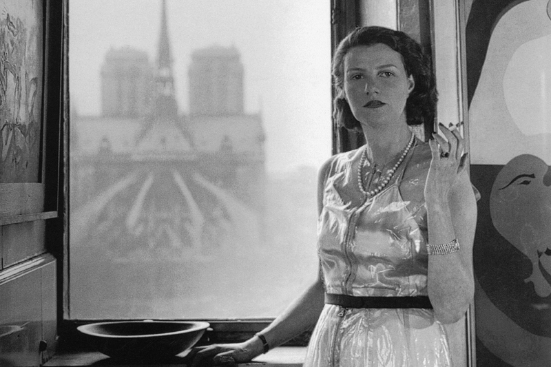 Peggy Guggenheim a Parigi fotografata da Rogi André, 1940. Foto in bianco e nero lei inpiedi davanti alla. Finestra con sigaretta. 