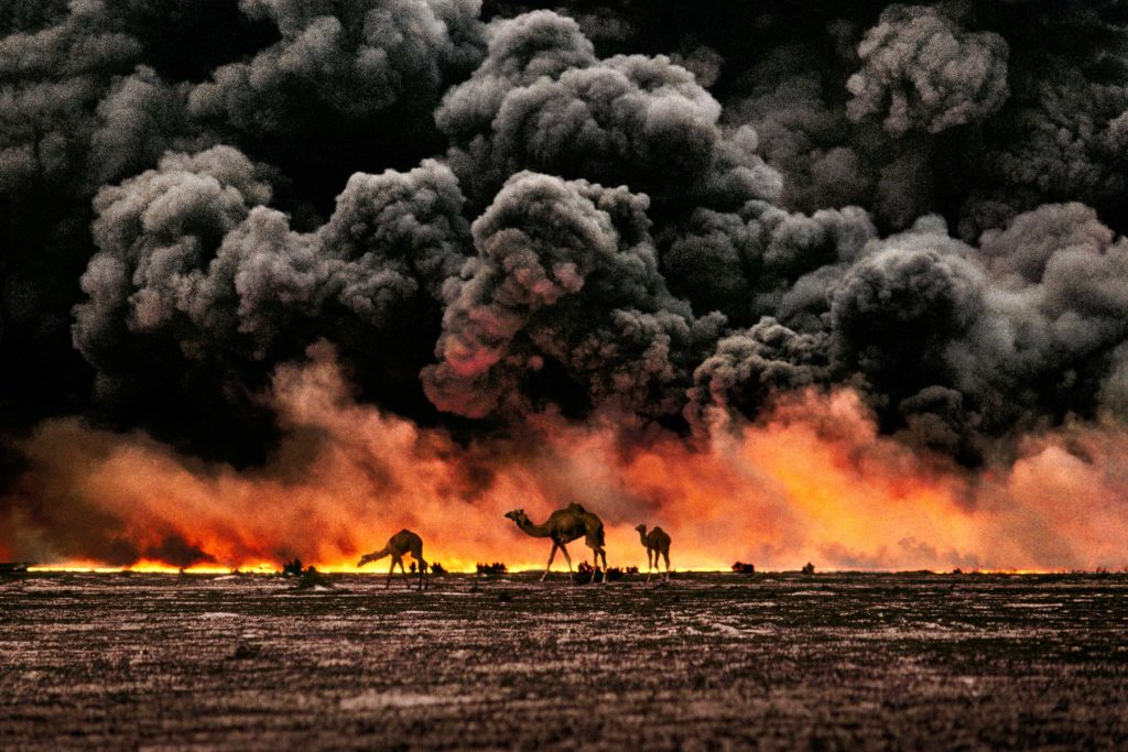 Scatto di Steve McCurry. Rappresenta dei cammelli mentre attraversano un pozzo di petrolio in fiamme. Sullo sfondo enormi nubi di fumo nero. 

