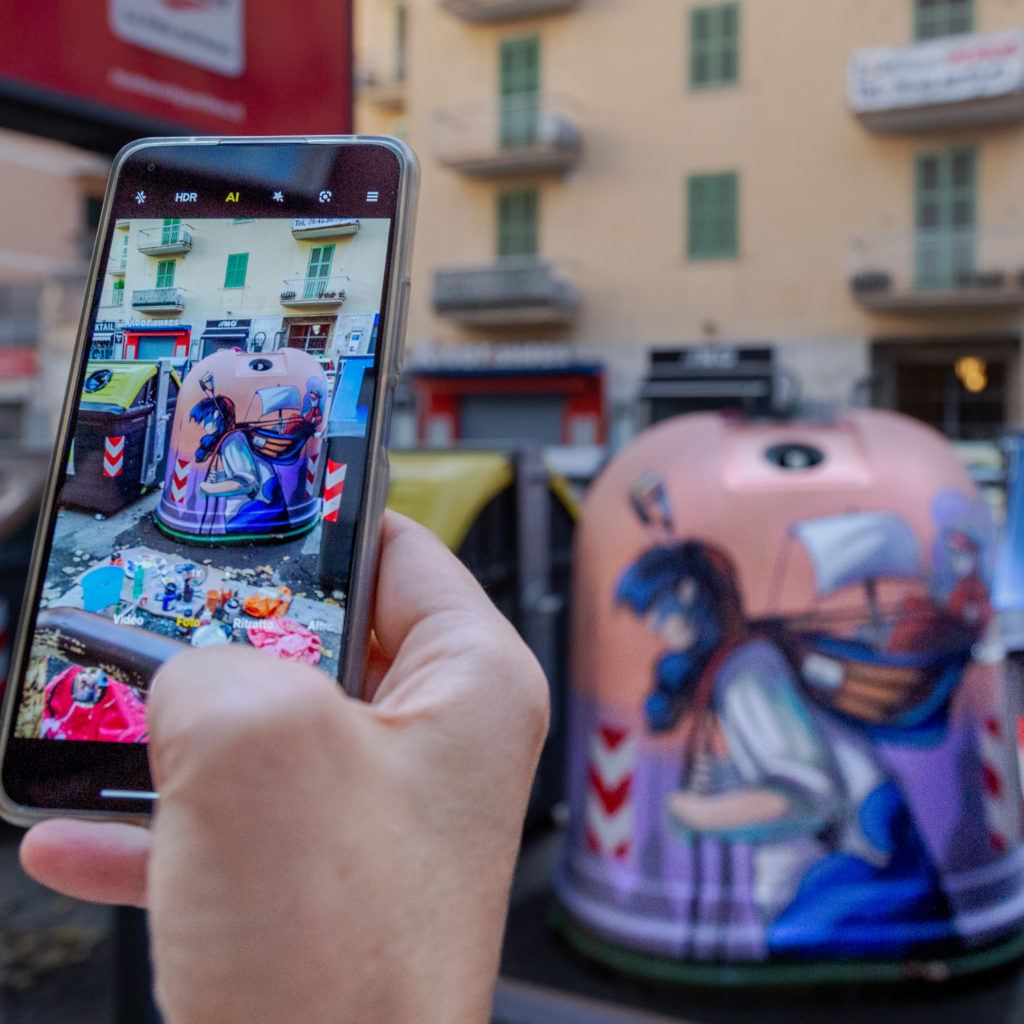 i CANTI DELL'INFERNO - UNA MANO IN PRIMO PIANO TIENE UN CELLULARE NELL'ATTO DI FOTOGRAFARE UNA CAMPANA DEL VETRO DIPINTA CON STREET ART, CON COLORE ROSA E DELLE FIGURE INFERNALI