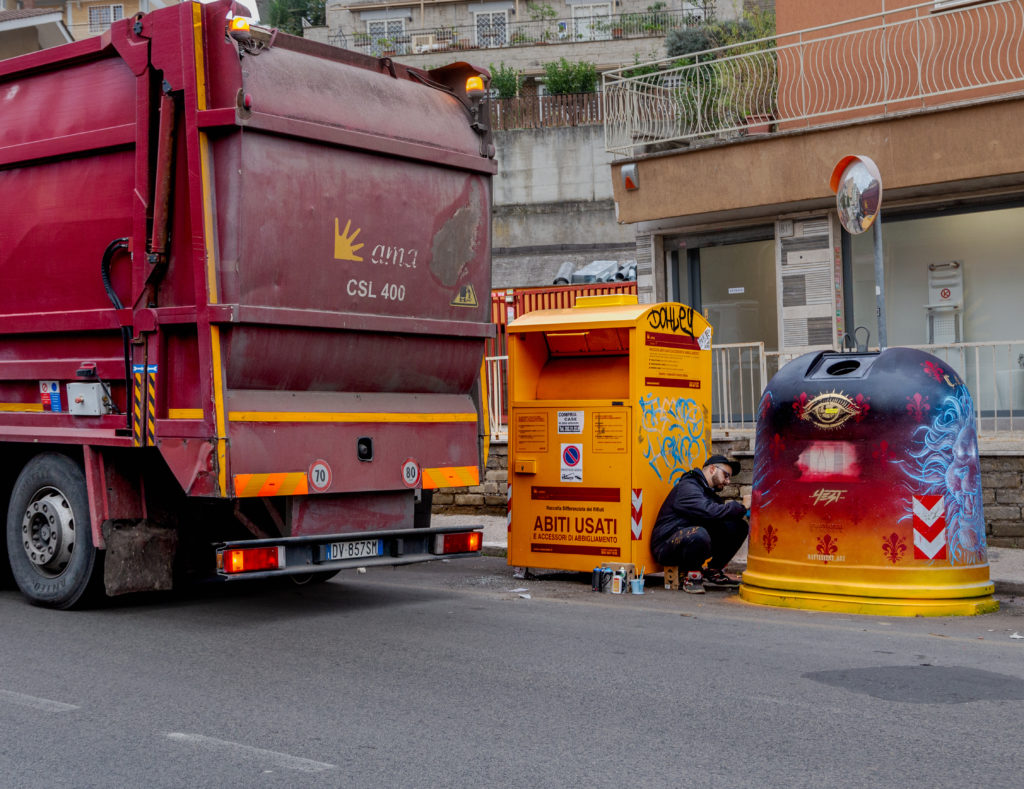 Inferno di Dante - Una campana del vetro disegnata con street art, in arancione con delle fiamme rosse, vicino un cassonetto dell'immondizia e un camion di raccolta rifiuti rosso della città di Roma