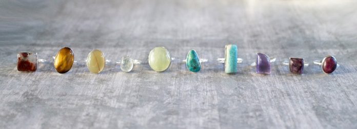 Cristalloterapia, antica proprietà, benessere, pietre. Dieci anelli con una pietra incastonata, tutte di colorazioni differenti, appoggiati su una superficie dura di colore grigio.