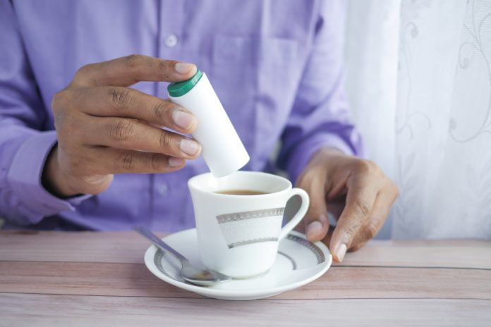 aspartame -un uomo vestito con camicia azzurra sta mettendo del dolcificante in una tazzina di caffè appoggiata su un piattino bianco con un cucchiaino a fianco