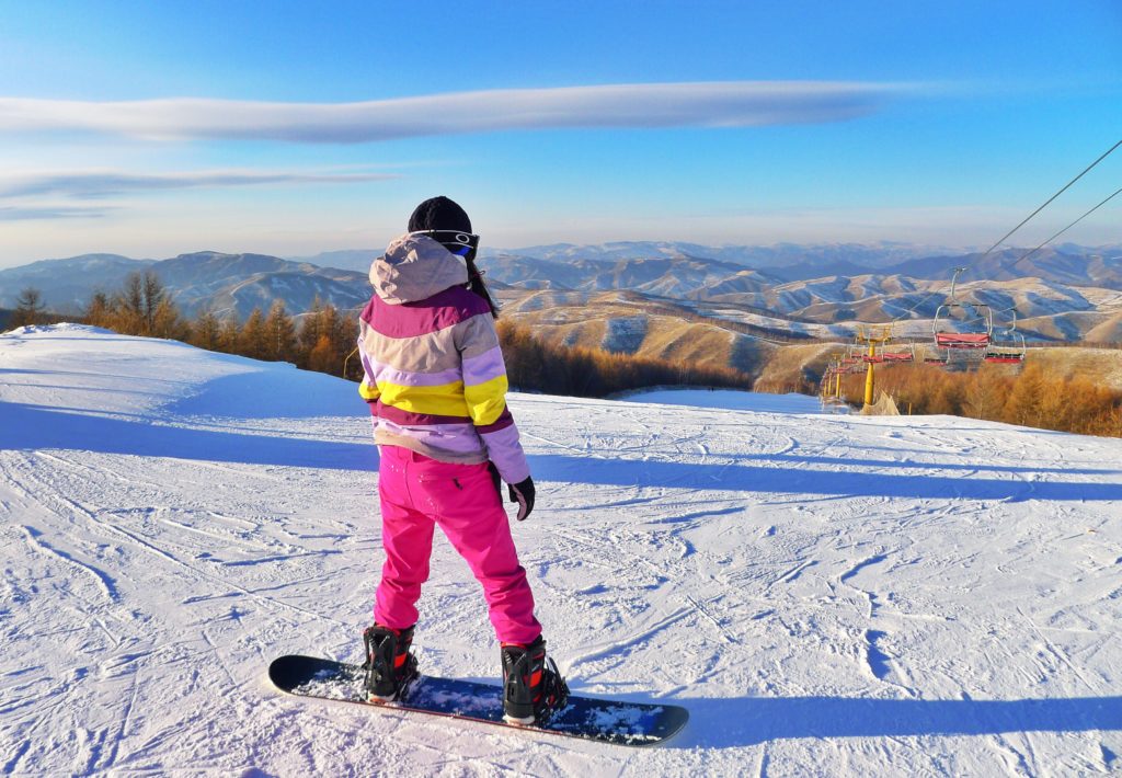 una ragazza con lo snowboard, su un apista da sci in discesa, la ragazza è di spalle e indossa una tuta bianca e rosa