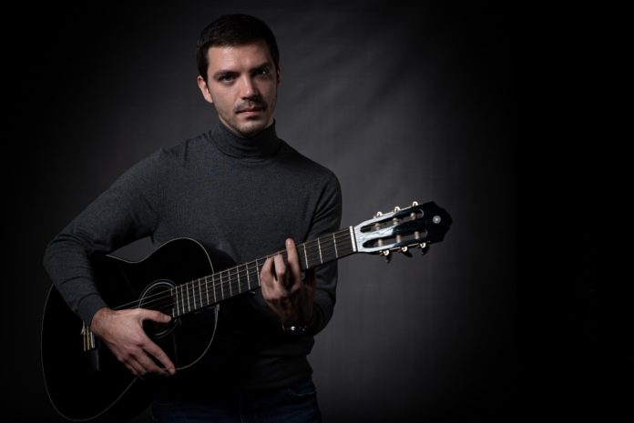 dario cuomo in primo piano, vestito di scuro, mentre imbraccia una chitarra acustica