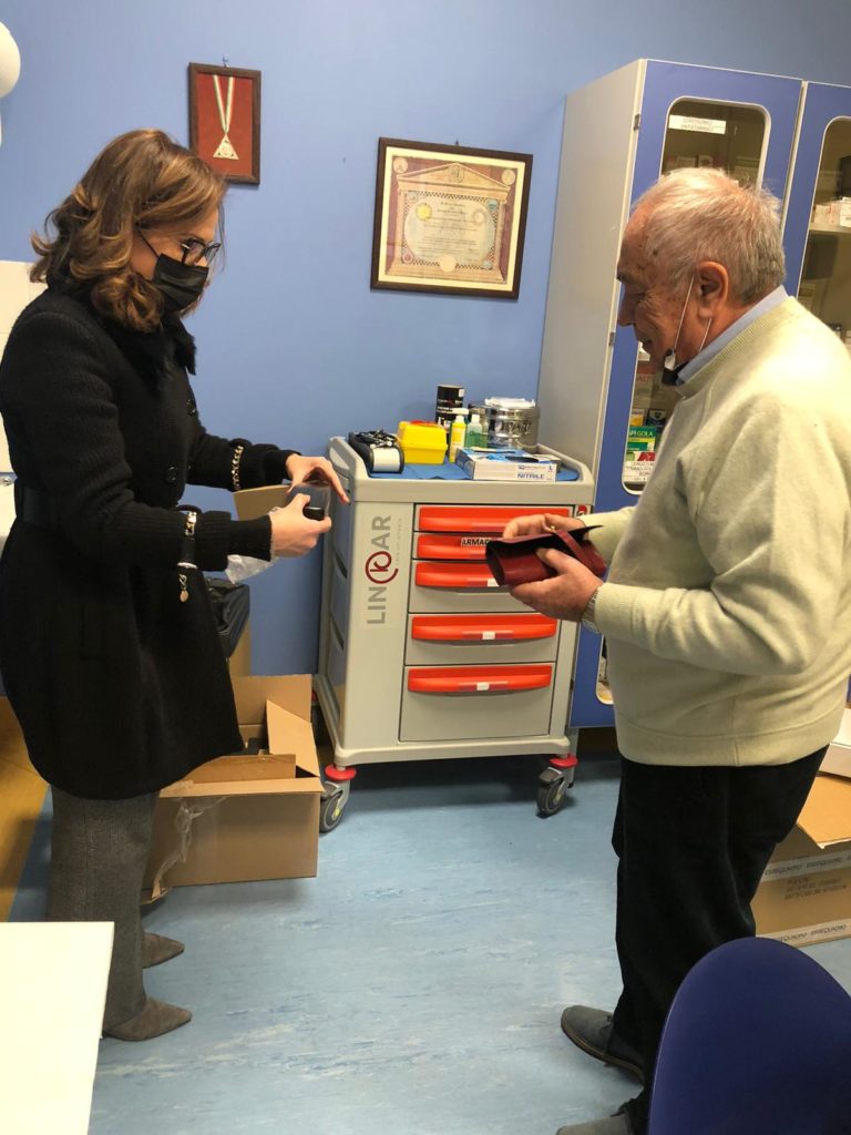 occhiali - nella foto una donna bionda ocn mascherina e capotto nero parla gesticolando conun uomo che indossa un maglione giallo chiaro e pantaloni scuri. I due sono in  un ambulatorio medico e si cvede una cassettiera con maniglie rosse e con sopra l'occorrente per medicazioni 