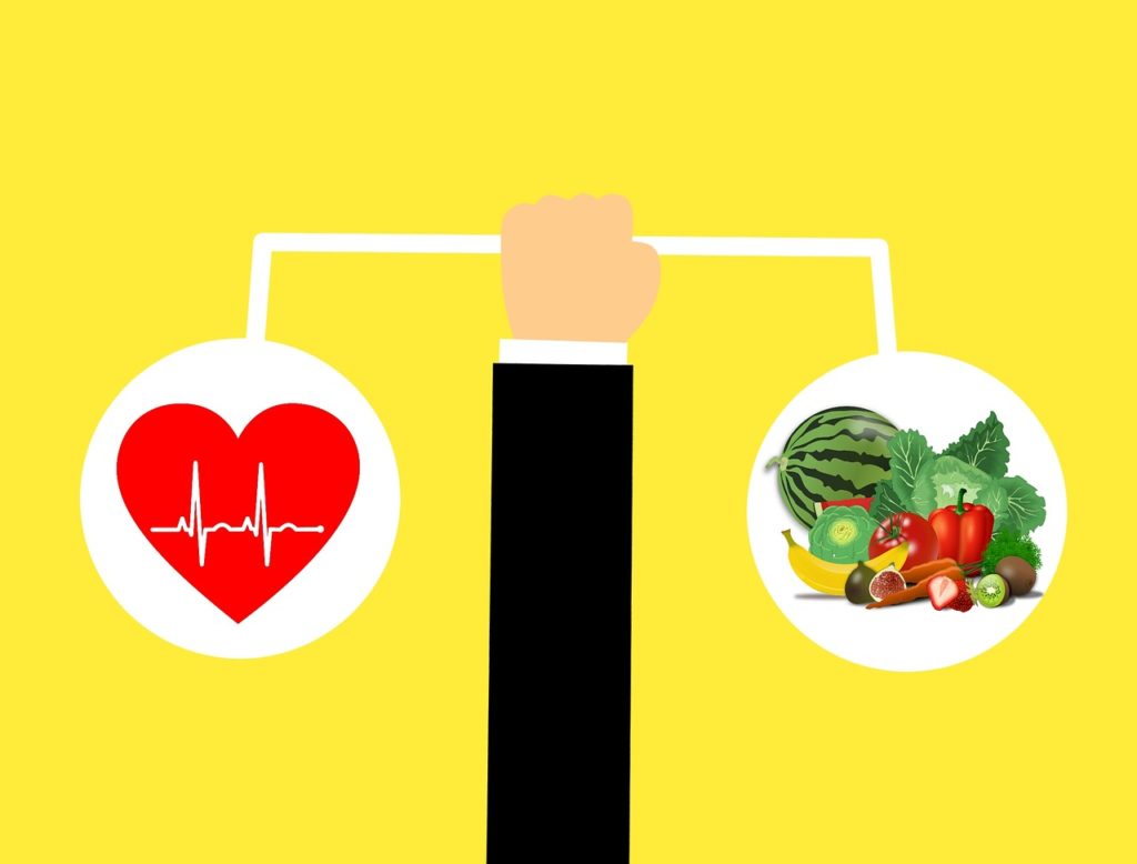 Un braccio che tiene una bilancia. Sulla sinistra un cuore rosso con sopra un disegno di un elettrocardiogramma e sulla destra un insieme di frutta e verdura. 