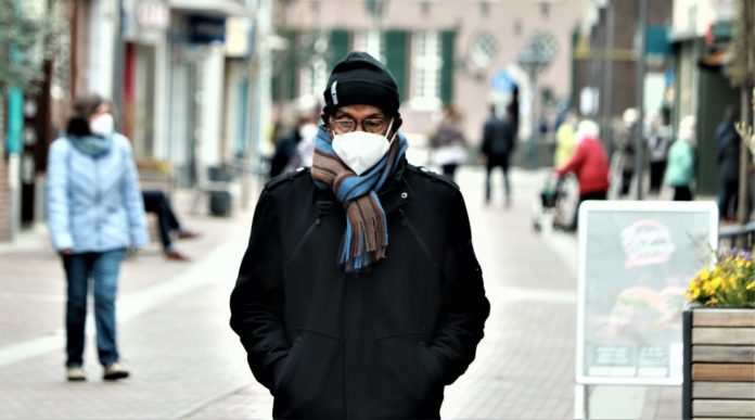 Mascherine all'aperto - un ragazzo vestito con cappotto scuro, sciarpa colorata intorno al collo, cappello di lana, occhiali, cammina in un avia del centro,con le mani in tasca. Intorno a lui alcuni passanti. Indossano tutti delle mascherine chirurgiche