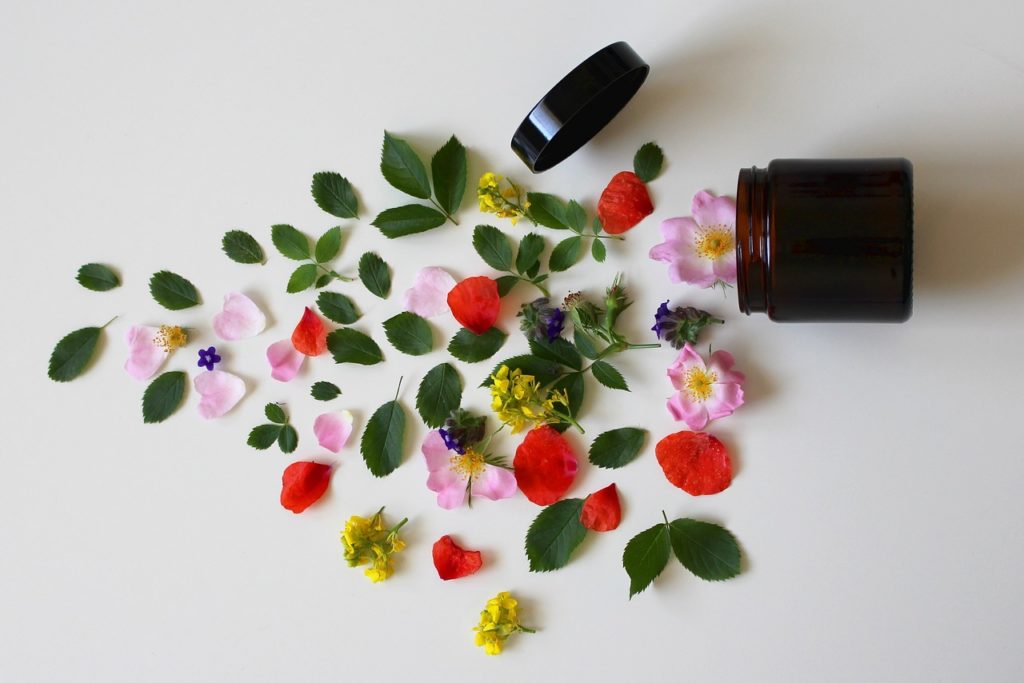 Un barattolo aperto da cui fuoriescono fiori rosa, petali rossi e piccole foglie di colore verde scuro. 
