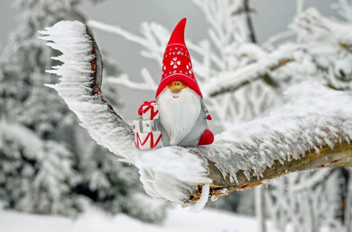 Babbo Natale rappresentato da una statuina appoggiata su dei rami pieni di neve. Indossa un cappuccio rosso ed ha una lunga barba bianca e con la mano destra tiene dei pacchetti natalizi