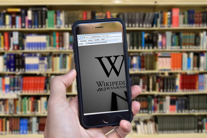wikipedia - nella foto la mano di un ragazzo tiene un telefonino con la pagina di wikipedia sullo schermo. Sullo sfondo, sfocata, una libreria piena di volumi e di libri
