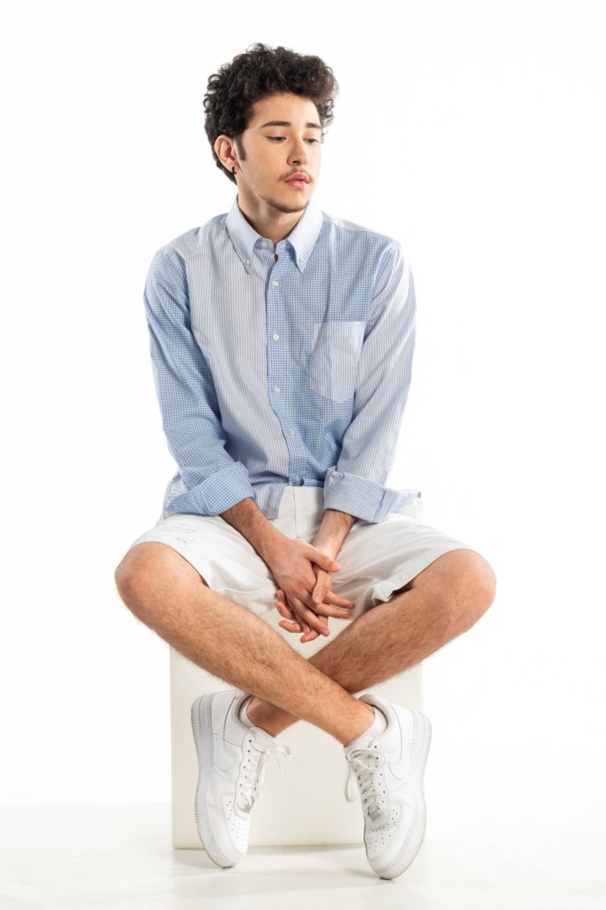 marcus, seduto su uno sgabello, indossa bermuda bianchi, camicia azzurra e scarpe da ginnastica bianche
