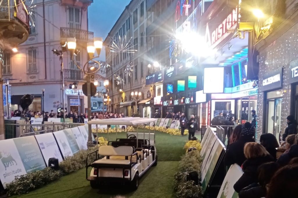 il red carpet di colore verde davanti al teatro ariston di sanremo, sul borso ci sono delle transenne con tanta gente appoggiata e in mezzo al red carpet c'è una macchinina elettrica scopderta