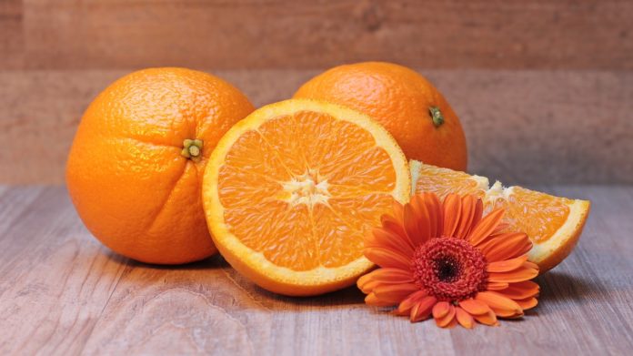 Vitamina C, proprietà, benefici, integratori alimentari. Tre arance, tra cui una tagliata a metà, poggiate su un tavolo in legno con un fiore di color arancione.