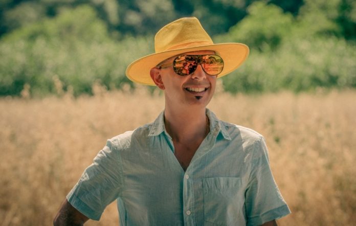 baldi giovani a sanremo #5 - robie c fotografao in un campo di grano, indossa una camicia azzurra, occhiali da sole e un cappello di paglia a tesa larga