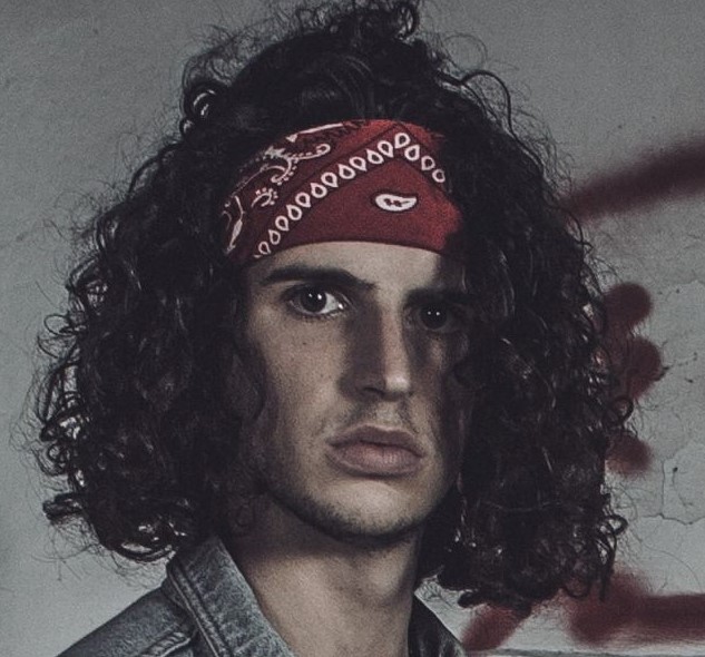 Guglielmo- Foto in stile vintage del cantautore. Riccio con i capelli lunghi indossa una giacca di jeans sopra una maglia nera scollata. Al collo porta una collana mentre i capelli sono tenuti indietro da una bandana rossa con una fantasia bianca.