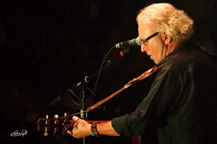 max brara di profilo, vestito di scuro, intento a cantare davanti al microfono, con la chitarra a tracolla