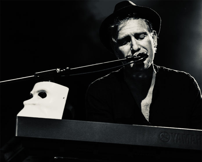 la droga - pianista indie in una foto in bianco e nero che lo ritrae intento a suonaare un piano elettrico, il microfono davanti alla bocca, una maschera bianca appoggiaataa sul bordo dello strumento