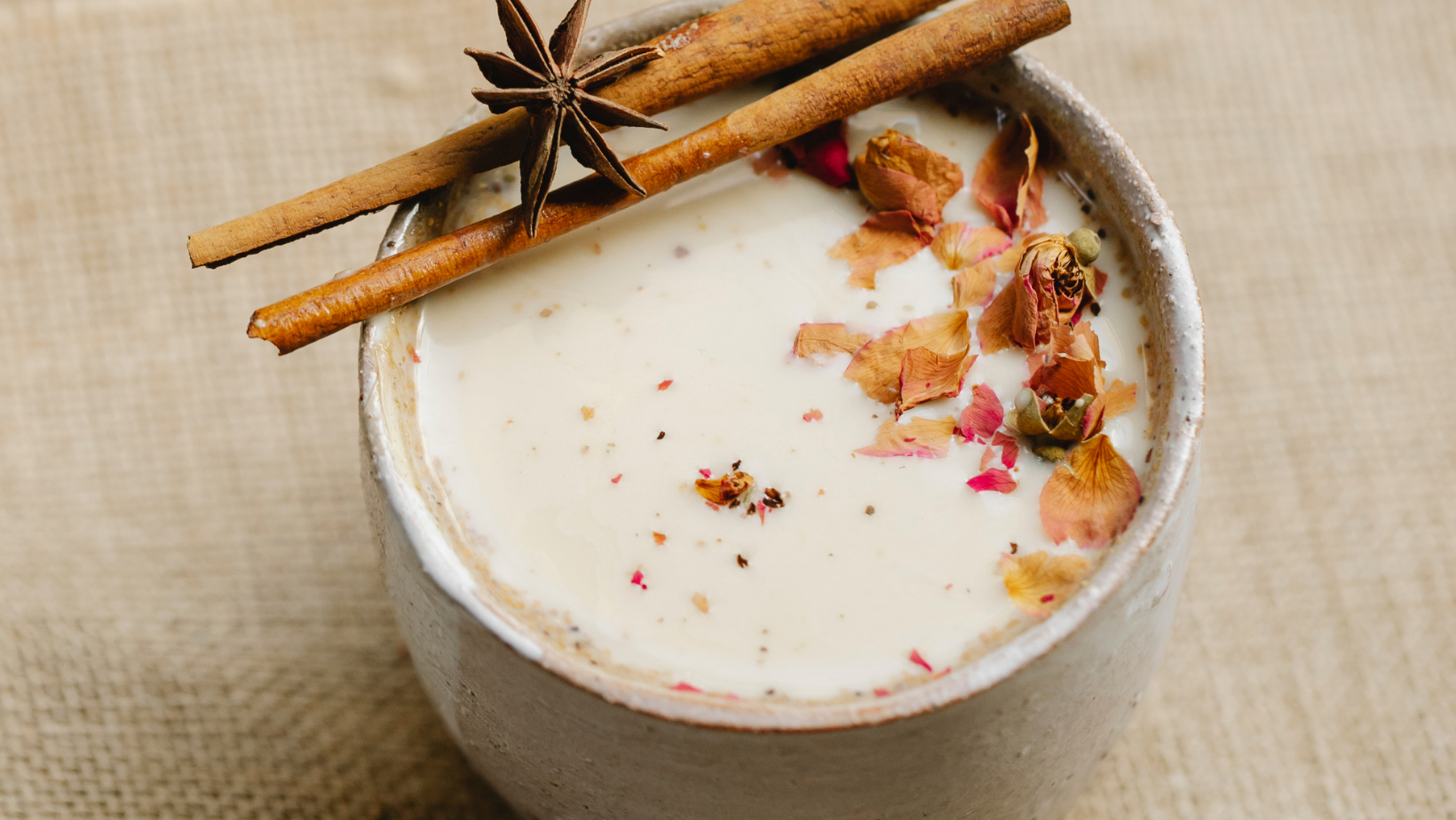 Nell'immagine c'è una tazzina bianca con all'interno il chai, il tipico tè indiano. Sulla superficie del liquido galleggiano delle spezie e sul bordo della tazzina sono appoggiate due stecche di cannella e un anice stellato.