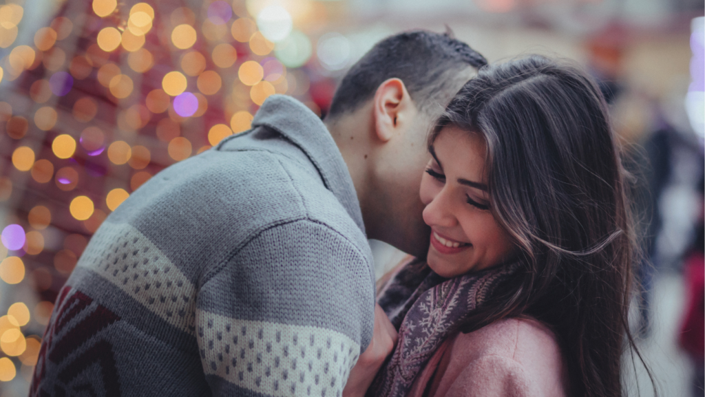 Un ragazzo dà un bacio sul collo alla partner. In sfondo le luci di natale.