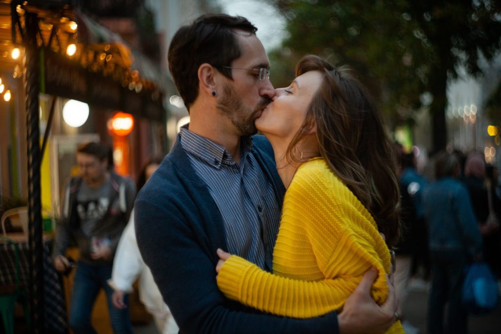 Bacio impacciato nella via di una città. La donna indossa un maglioncino giallo mentre l'uomo una camicia a righe bianche e blu con sopra un cardigan blu.