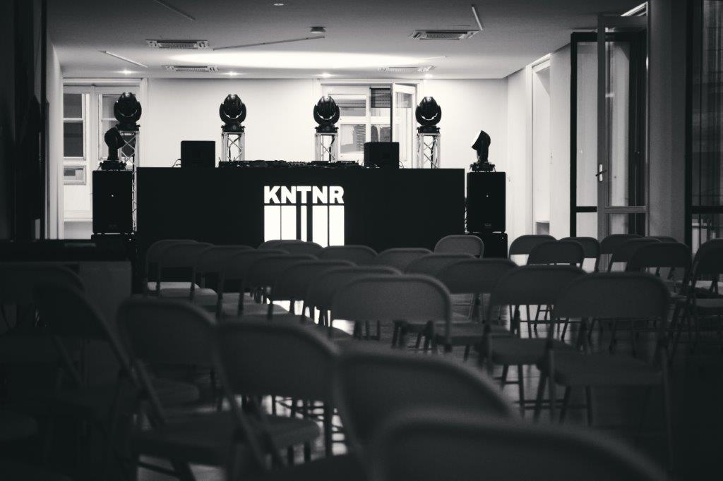 foto in bianco e nero di una location costituita da sedie e un bancone con la scritta KNTNR