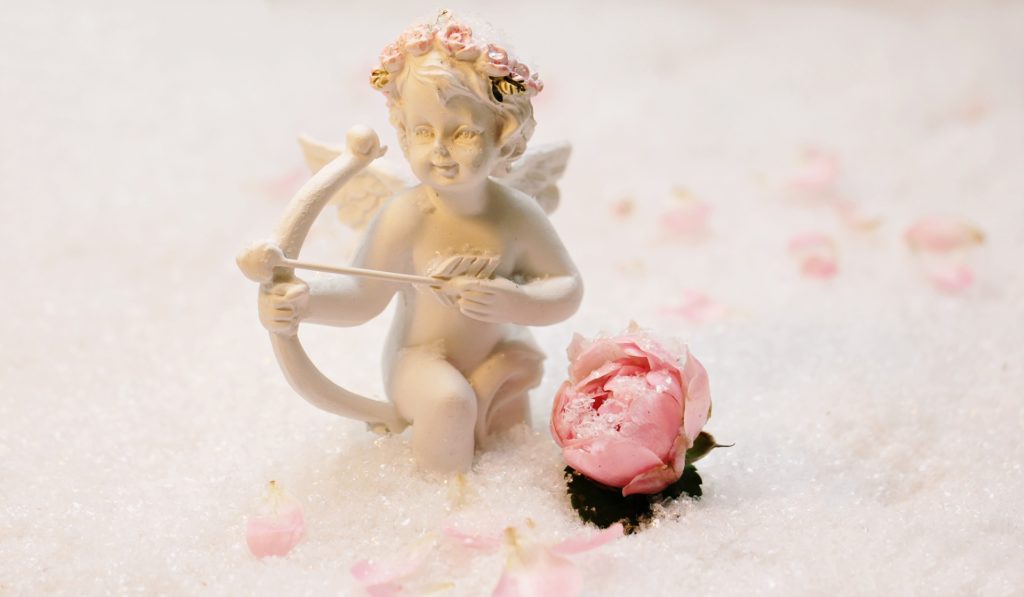 San Valentino Day - Un cupido di porcellana nell'intento di scagliare una freccia dal suo arco, indossa una corona di fiori. Vixino a lui c' è il bocciolo di una rosa rosa, e la statuina è appoggiata su petali di fiori