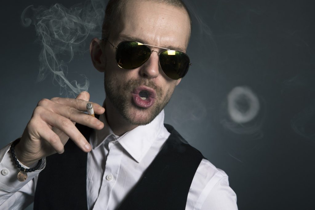 un uomo con camicia bianca e gillè nero, insodda occhiali da sole neri a goccia, mentre sbuffa una fumata di sigaretta con la bocca aperta, tra le dita tiene una sigaretta nera e ha un'aria superba e strafottente