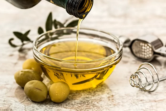 Olio di oliva, proprietà, cosmetica, rimedi naturali. Una ciotolina di vetro piena di olio con la bottiglia e delle olive verdi intorno.