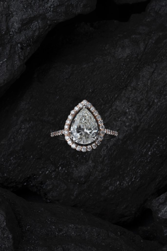 JGT- Anello con grande diamante centrale a goccia contornato di piccoli diamanti (microincassatura). Lo sfondo è costituito da una roccia nera che risalta la lucentezza del diamante.
